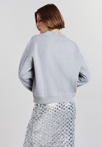 Donna con i capelli corti castani, che indossa un maglione grigio chiaro e una gonna di paillettes argento, voltata leggermente di lato su uno sfondo semplice.