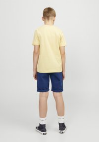 Gul bomull t-skjorte, blå rullede shorts, hvite sokker og marineblå sko. Utsikt fra bak, som viser det enkle designet og fargekontrasten.