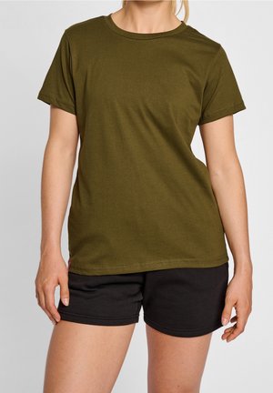 Osoba ubrana w oliwkowozieloną koszulkę z krótkim rękawem i czarne szorty, stojąca na tle jasnego, jednolitego tła.