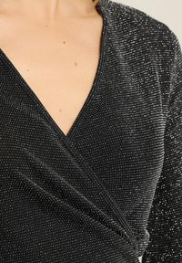 Primo piano di una blusa avvolgente nera e scintillante con scollo a V indossata da una persona, che mostra la texture del tessuto e il décolleté.