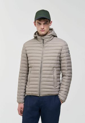 Jonge man draagt een lichtgrijze doorgestikte jas met capuchon en marineblauwe broek, staand tegen een effen lichte achtergrond, handen in de zakken.