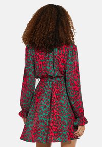 Kurzes Langarmkleid mit strukturiertem Schnitt. Es hat ein lebhaftes rotes und grünes Tiermuster, eine taillierte Taille und Rüschennähte an den Ärmeln.
