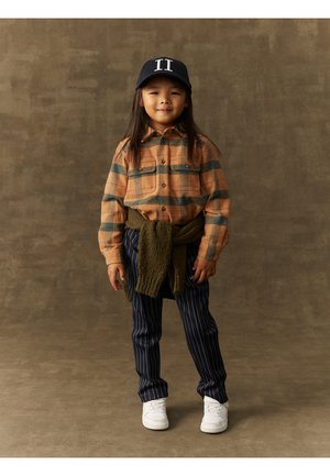 Rutete flanellskjorte i oransje og grønn, olivengrønn genser knyttet rundt livet, marineblå stripete bukser, hvite sneakers, og marineblå caps med "II" detalj.