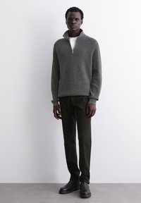 Grauer gerippter Strickpullover mit einem Reißverschlusskragen, kombiniert mit dunkelgrünen Hosen und schwarzen Knöchelschuhen vor einem neutralen Hintergrund.