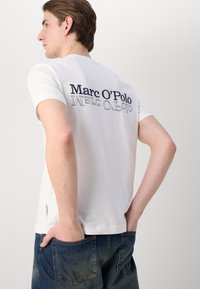 Hvid bomulds T-shirt med "Marc O'Polo" logo i marineblå på ryggen. Korte ærmer, rund halsudskæring og afslappet pasform. Enkel design.