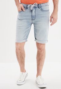 Shorts en denim bleu clair avec des revers, dotés d'une taille élastique avec cordon de serrage et de poches avant. Portés avec des baskets blanches.