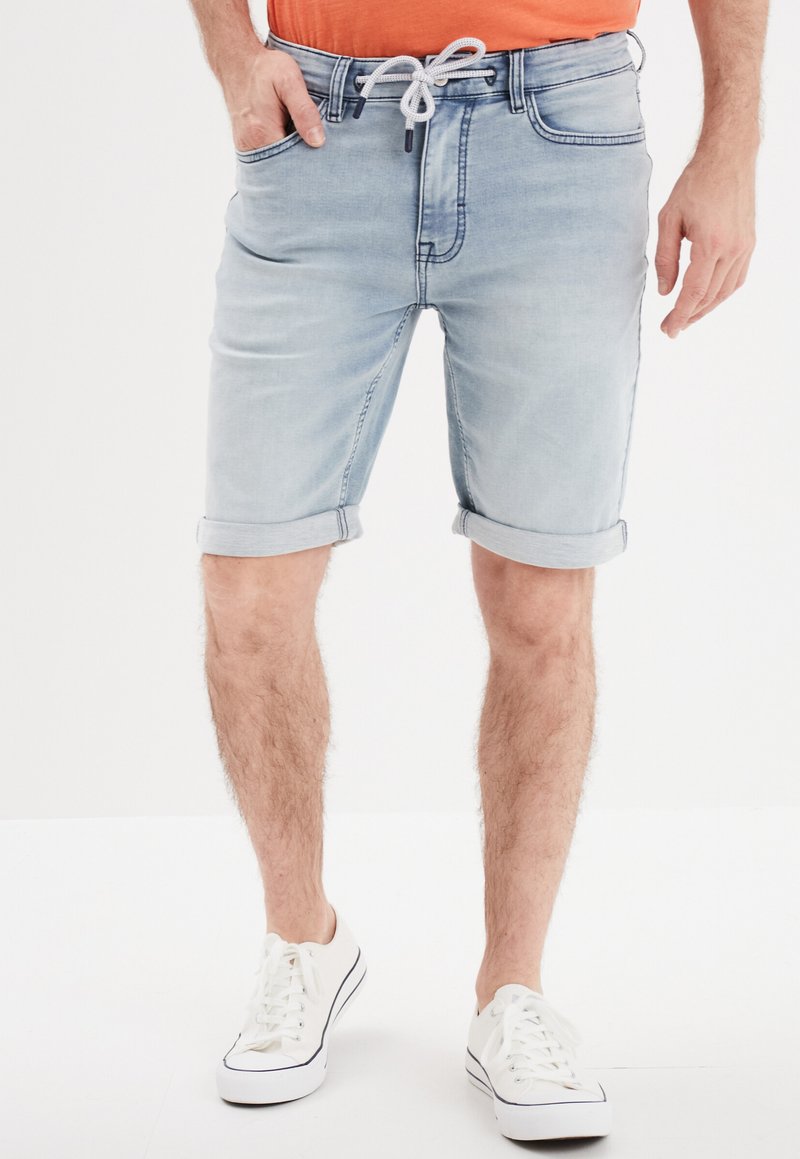 Shorts en denim bleu clair avec des revers, dotés d'une taille élastique avec cordon de serrage et de poches avant. Portés avec des baskets blanches.