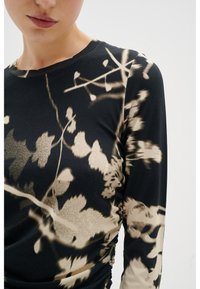 Mujer vistiendo una camiseta de manga larga negra con patrones florales abstractos en beige, mostrada desde el cuello hasta la cintura contra un fondo liso.