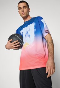 Maillot de basketball en maille multicolore présentant un dégradé de bleu à rose, des manches courtes et un design imprimé. Un ballon de basket noir tenu dans une main.