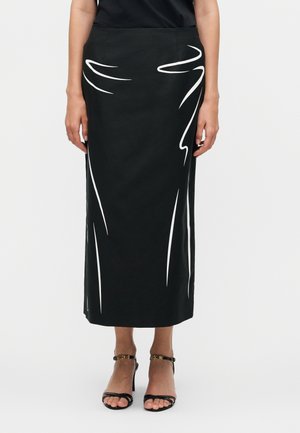 Kobieta nosząca czarną midi spódnicę z białymi abstrakcyjnymi wzorami linii, zestawioną z czarnymi sandałami na wysokim obcasie z paskami wokół kostki.