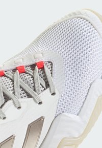 Zapato deportivo con parte superior de malla blanca, acentos plateados texturizados, cordones grises y lazos de cordón de color coral brillante, con una suela de goma firme.