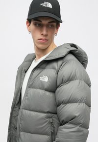 Szürke puffertakaró kapucnival, steppelt mintával és logóval. Fehér ing fölött viselve, fekete sapkával, amelyen logó található.