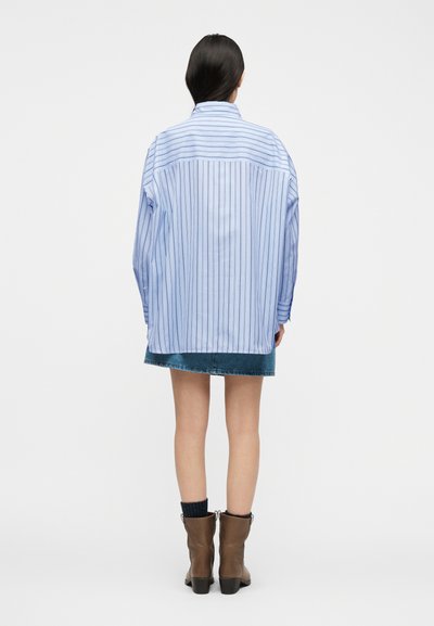 Γυναίκα με μακριά σκούρα μαλλιά, ντυμένη με υπερμεγέθη ανοιχτό μπλε ριγέ πουκάμισο, τζιν φούστα, μαύρες κάλτσες και καφέ μποτάκια μέχρι τον αστράγαλο, γυρισμένη από την πίσω πλευρά.