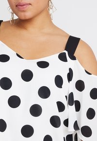 Top nero a pois su sfondo bianco, con design a spalle scoperte, ampia scollatura e spalline nere. Tessuto morbido e strutturato.