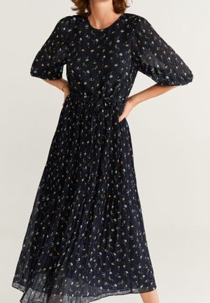Frau trägt ein langes schwarzes Kleid mit kleinen Blumenmustern, Puffärmeln und einem gebundenen Taillengürtel, steht mit den Händen in der Hüfte.