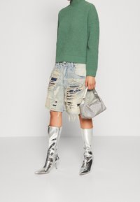 Pull à col roulé en maille verte, jupe en denim bleu clair déchiré, bottes argentées à talons hauts, et un petit sac à main métallique.