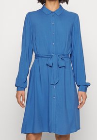 Abito blu con bottoni, colletto, maniche lunghe e vita legata. Tessuto morbido con una texture liscia e una leggera lucentezza, che termina sopra il ginocchio.