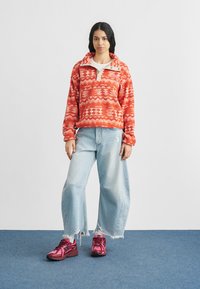 Νεαρή γυναίκα στέκεται φορώντας κόκκινο πουλόβερ με σχέδια, ανοιχτόχρωμα τζιν με φαρδιά σκισμένα πόδια και ροζ αθλητικά παπούτσια σε μπλε χαλί.