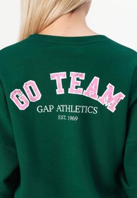 Sweatshirt vert avec "GO TEAM" en lettres roses usées, accompagné de "GAP ATHLETICS" et "EST. 1969" en blanc. Matériau doux, design col rond.