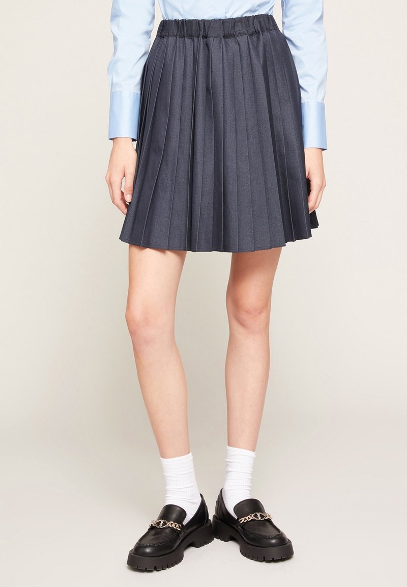 Personne portant une jupe plissée foncée, une chemise bleu clair à manches longues, des chaussettes blanches et des mocassins noirs avec un détail de chaîne dorée, debout sur un fond uni.