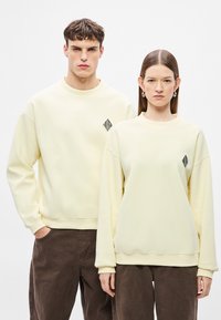 Geltoni megzti džemperiai su juodu deimanto formos logotipu krūtinėje. Modelio kelnės tamsiai rudos spalvos. Audinys atrodo minkštas ir šiek tiek tekstūruotas.