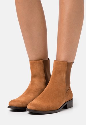 Bottines en daim marron avec panneaux élastiques sur les côtés et talons bas empilés, portées sur jambes nues contre un fond blanc.