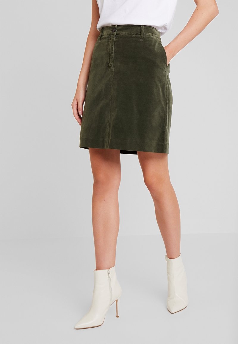 Femme portant une jupe en velours côtelé vert olive, un haut blanc et des bottines blanches à talons, debout avec une main dans la poche.