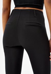 Nära bild av en person som bär högmidjade svarta tajta byxor med bakfickor och en kortärmad stickad svart topp.