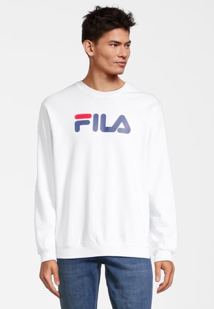 Felpa bianca con maniche lunghe che presenta il logo Fila in rosso e blu. Tessuto morbido, polsini e orlo a coste, vestibilità comoda. Jeans blu indossati sotto.
