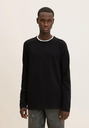 Schwarz gewebtes Langarmshirt mit weißem Rundhalsausschnitt und Raglanärmeln, kombiniert mit grauen verwaschenen Jeans.