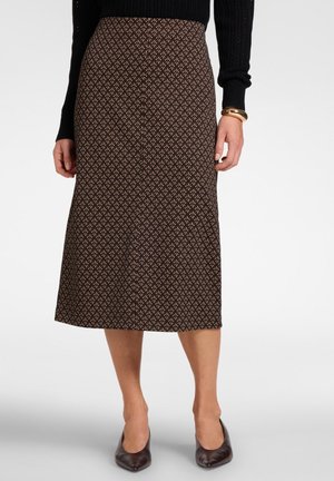 Donna che indossa una gonna midi marrone a fantasia, top nero a maniche lunghe, braccialetti dorati e scarpe marrone scuro con punta appuntita, in piedi su uno sfondo semplice.
