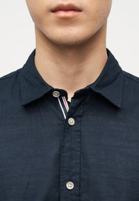 Κοντινή λήψη ενός ατόμου που φοράει σκούρο μπλε πουκάμισο με κουμπιά και γιακά, εμφανίζοντας μέρος του λαιμού και του κάτω μέρους του προσώπου.