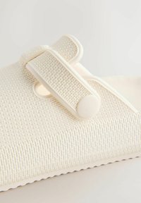 Witte, gestructureerde instapschoen met een brede band en drukknopsluiting. Heeft een subtiel geribd oppervlak en een gevormde, flexibele vorm.