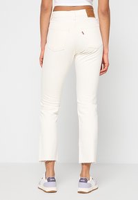 Jeans de mezclilla blanca con dobladillo deshilachado, corte ajustado y bolsillos traseros. Presenta un parche de cuero marrón en la cintura. Llevados con zapatillas blancas.