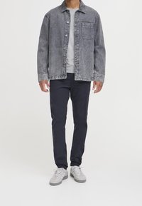 Giacca di jeans in grigio sbiadito con tasca frontale, abbinata a pantaloni blu scuro e sneakers grigio chiaro. Design semplice e strutturato.