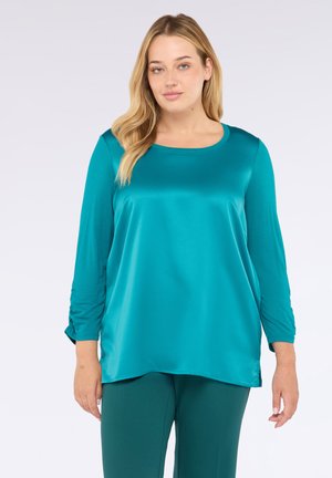 Donna bionda che indossa una blusa di raso satinato a maniche lunghe color turchese e pantaloni abbinati, su uno sfondo grigio chiaro uniforme.