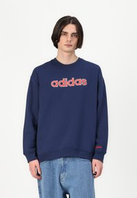 Tamnoplavi dukserica s crvenim "adidas" logotipom na prsima, okrugli izrez i duge rukave. Ima rebraste manžete i donji rub.