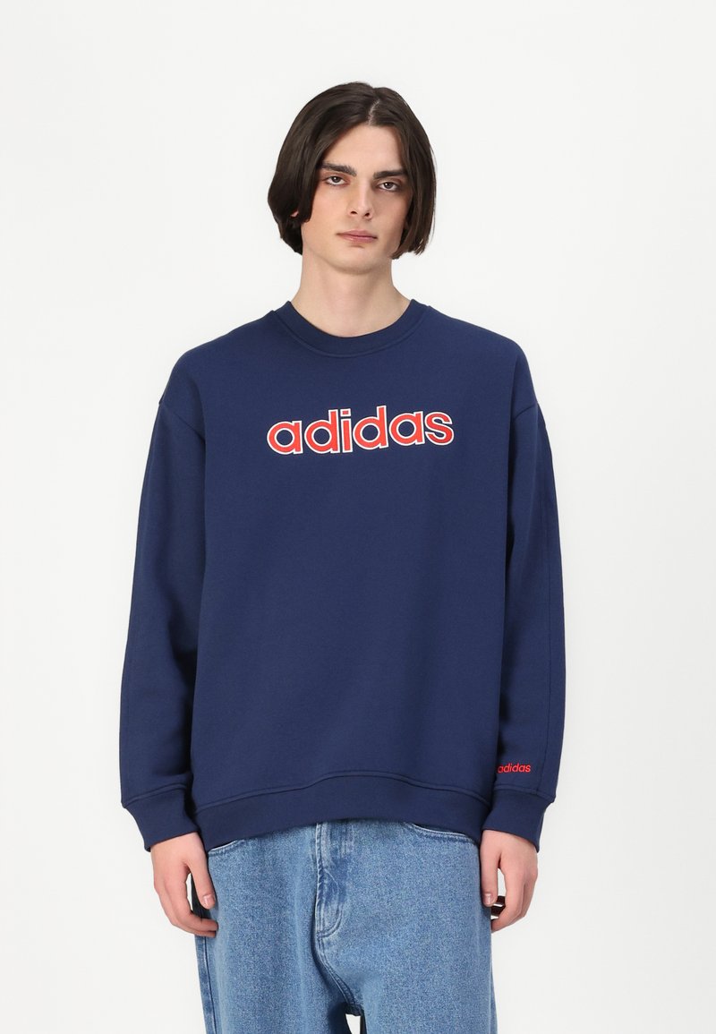 Tamnoplavi dukserica s crvenim "adidas" logotipom na prsima, okrugli izrez i duge rukave. Ima rebraste manžete i donji rub.