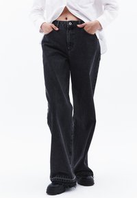 Pantalones de mezclilla anchos negros con cintura alta, que presentan bolsillos delanteros y un dobladillo deshilachado, combinados con calzado negro grueso.