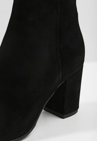 Bottine en suede noire avec un bout arrondi et un talon bloc épais. Texture lisse avec une finition haute et sans quincaillerie visible.