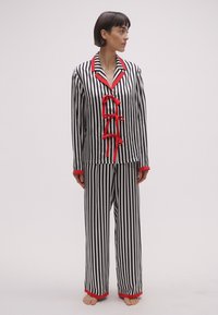 Set de pijama cu dungi alb-negru și accente roșii, având un top cu nasturi, trei panglici decorative și pantaloni largi asortați.