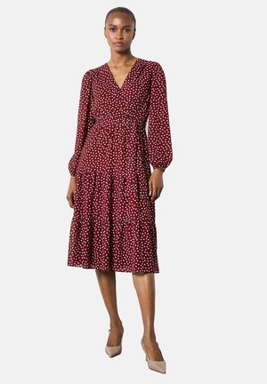 Donna in un abito midi a maniche lunghe bordeaux con pois bianchi e décolleté beige a punta su uno sfondo bianco.