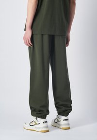 Groene stoffen sweatpants met elastische boorden, relaxte pasvorm. Gecombineerd met witte sneakers met zwarte en gele accenten, gezien vanuit de achterkant.