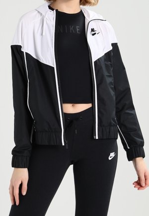 Woman wearing black and white Nike tracksuit with a cropped top, zip-up jacket, and leggings, showing midriff and brand logos.