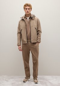 Giacca impermeabile beige con cappuccio, tasche con zip; maglione fitted beige sotto. Pantaloni beige abbinati e sneakers con dettagli.