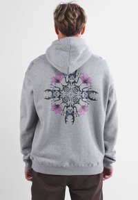 Persona che indossa un hoodie grigio con un design simmetrico sulla schiena di scarafaggi cervo neri e fiori rosa, in piedi su uno sfondo neutro.