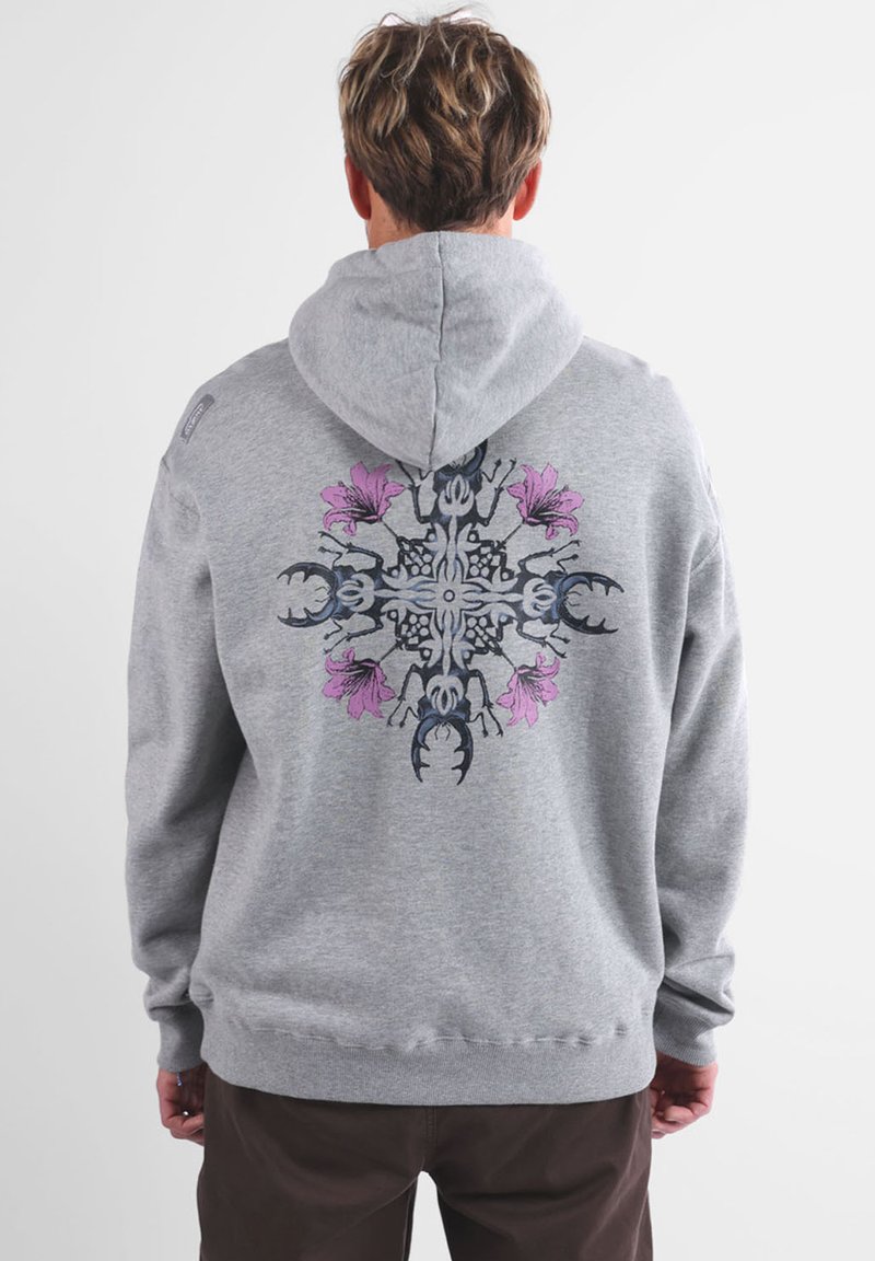 Persona che indossa un hoodie grigio con un design simmetrico sulla schiena di scarafaggi cervo neri e fiori rosa, in piedi su uno sfondo neutro.