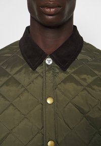 Olivgrön quiltad jacka med mörkbrun kordfläskkrage, utrustad med mässingsknappar och diamanterställda sömmar.