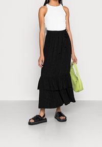 Jupe maxi noire à volants avec un design texturé, associée à un débardeur blanc et des sandales compensées noires. Sac vert à la main.