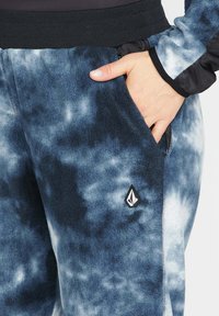Blaue Batik-Fleecehosen mit einem schwarzen elastischen Taillengummi, Reißverschlusstaschen und einer strukturierten Oberfläche, die ein Logo-Detail aufweisen.